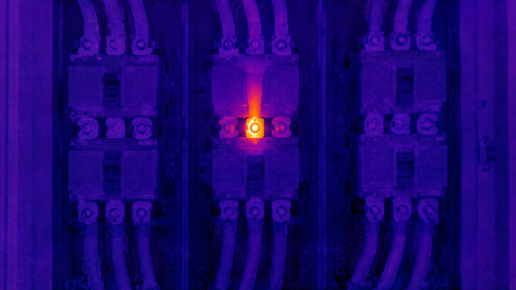Switchgear seen in thermal: three breakers in cool violet, one connection glowing orange with heat.